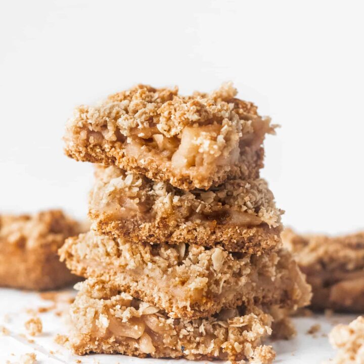 4 healthy apple crisp bars stacked on top of each other on parchment paper with more apple crisp in the background