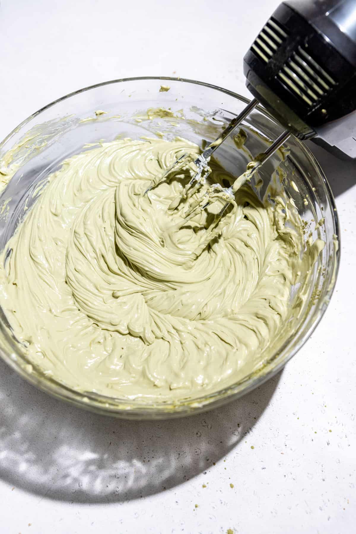 Matcha cheesecake filling being mixed together by a hand mixer in a glass bowl.