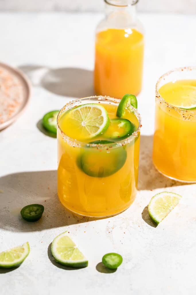 A short glass filled with orange margarita, garnished with jalapeño and lime slices with a sugared rim.