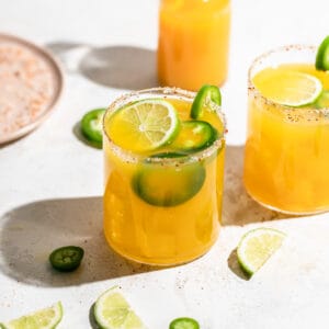 A short glass filled with spicy orange margarita, garnished with jalapeño and lime slices.