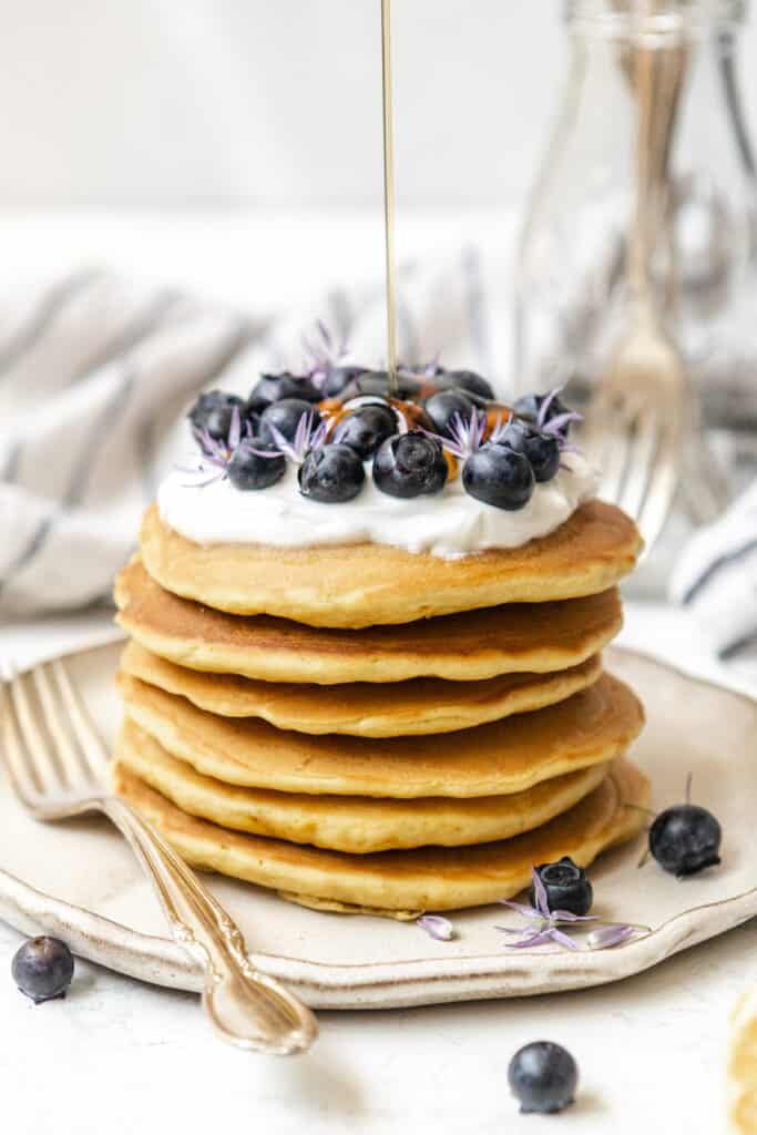 Maple syrup being poured on a stack of pancakes with blueberries.
