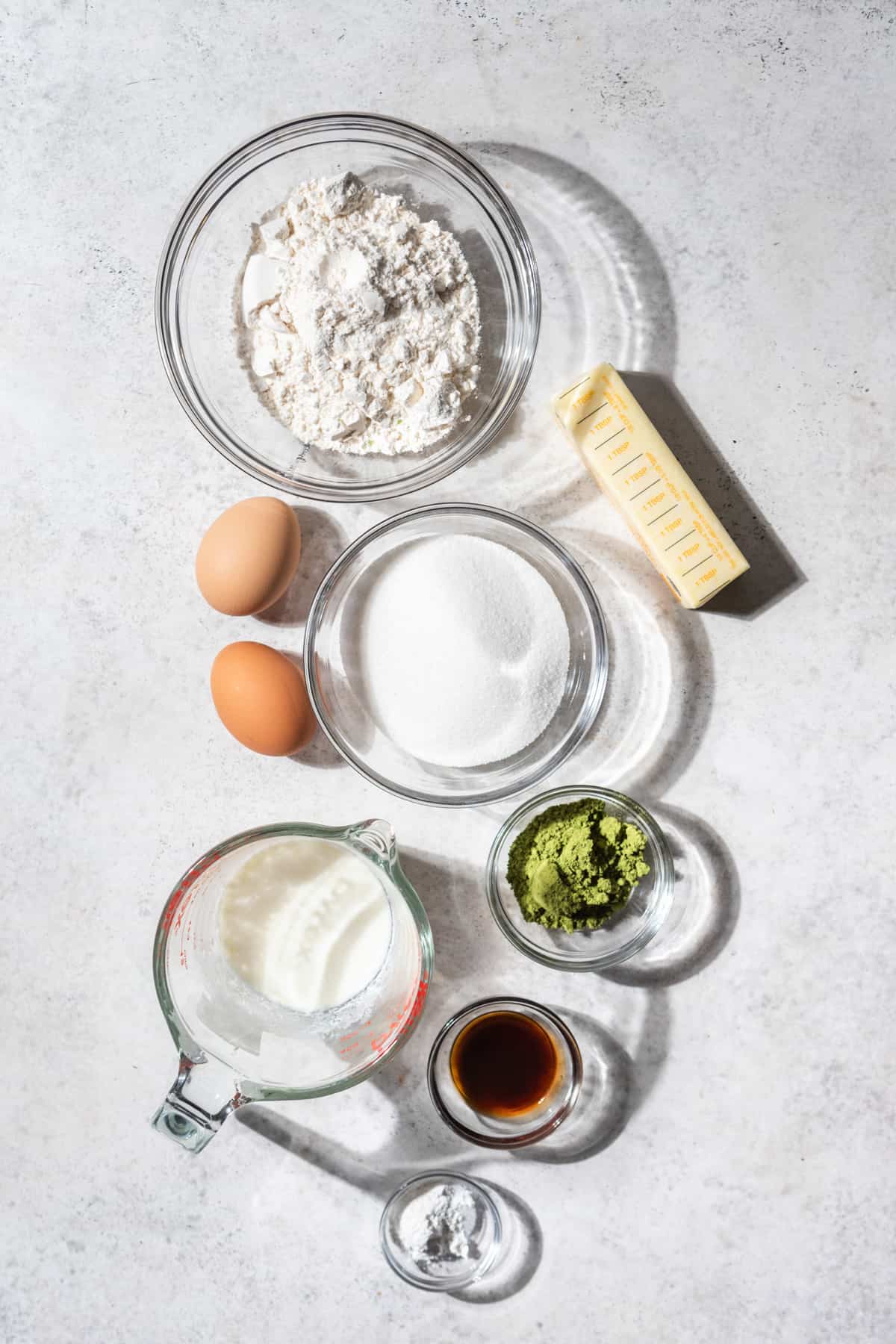 Bowls of flour, butter, sugar, matcha, buttermilk, vanilla, baking powder, and eggs on a white surface.