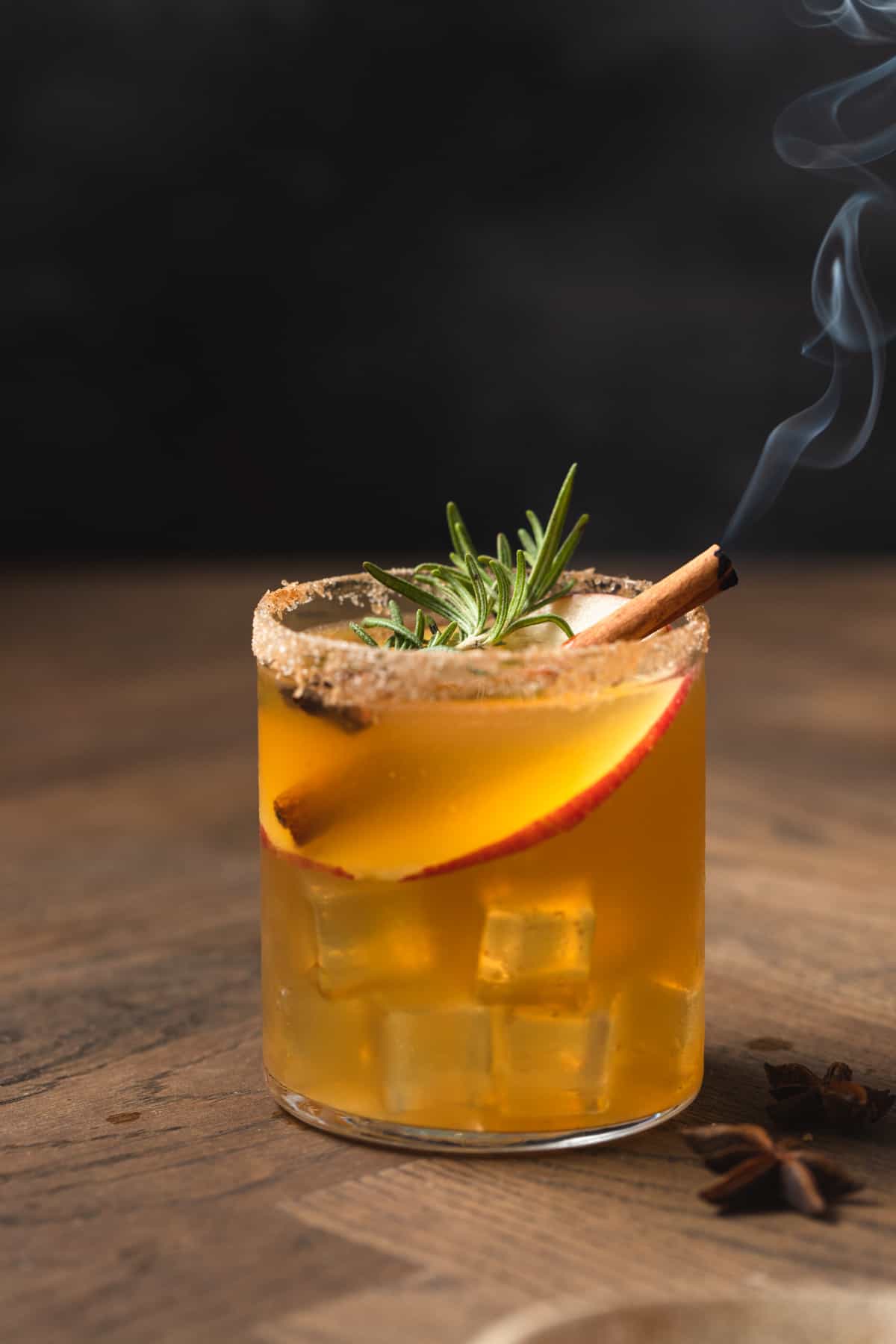 An orange cocktail garnished with an apple slice, rosemary, and a smoking cinnamon stick.