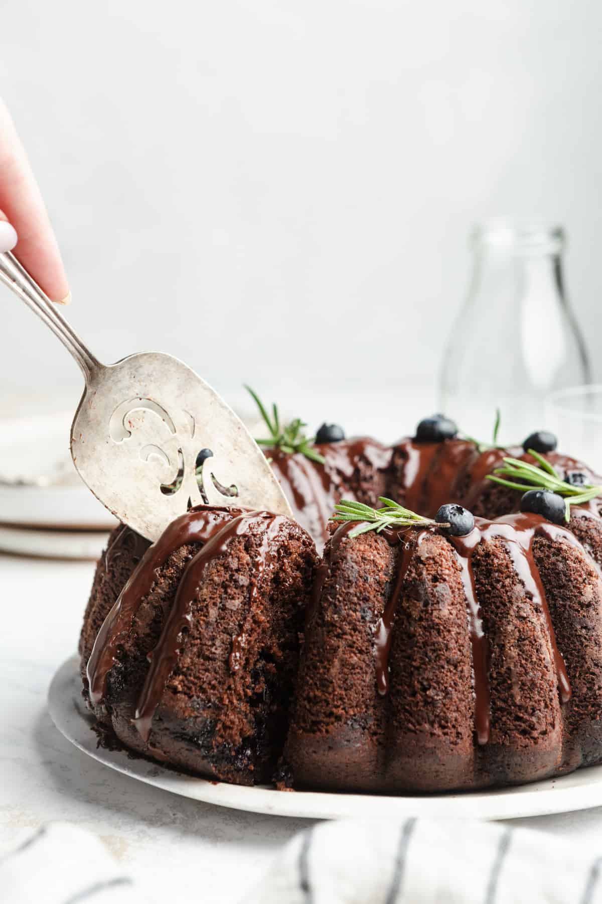 Slicing a chocolate bundt cake topped with blueberries and rosemary with a cake server.
