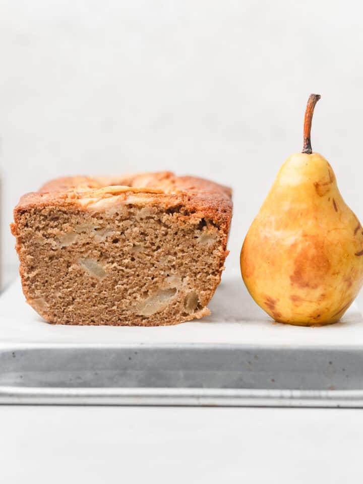 A loaf of vanilla pear bread next to a pear on an upside down baking sheet.