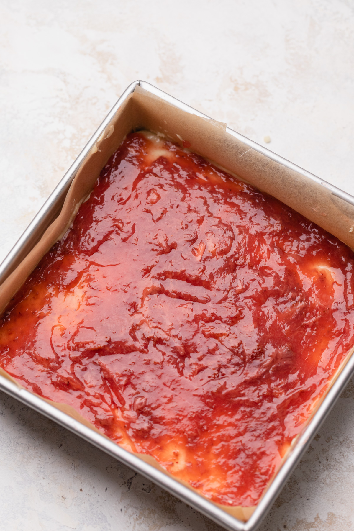 Strawberry jam spread over cake batter in a square pan.