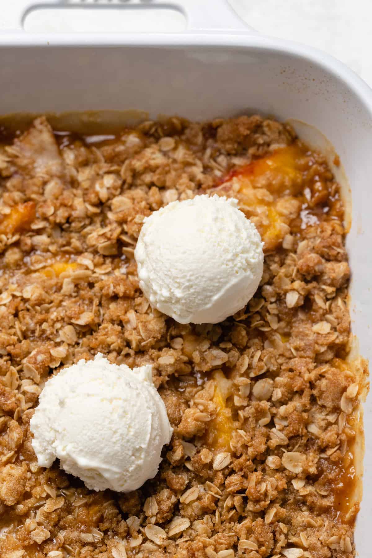 Peach crisp in a large dish with 2 scoops of vanilla ice cream on top.