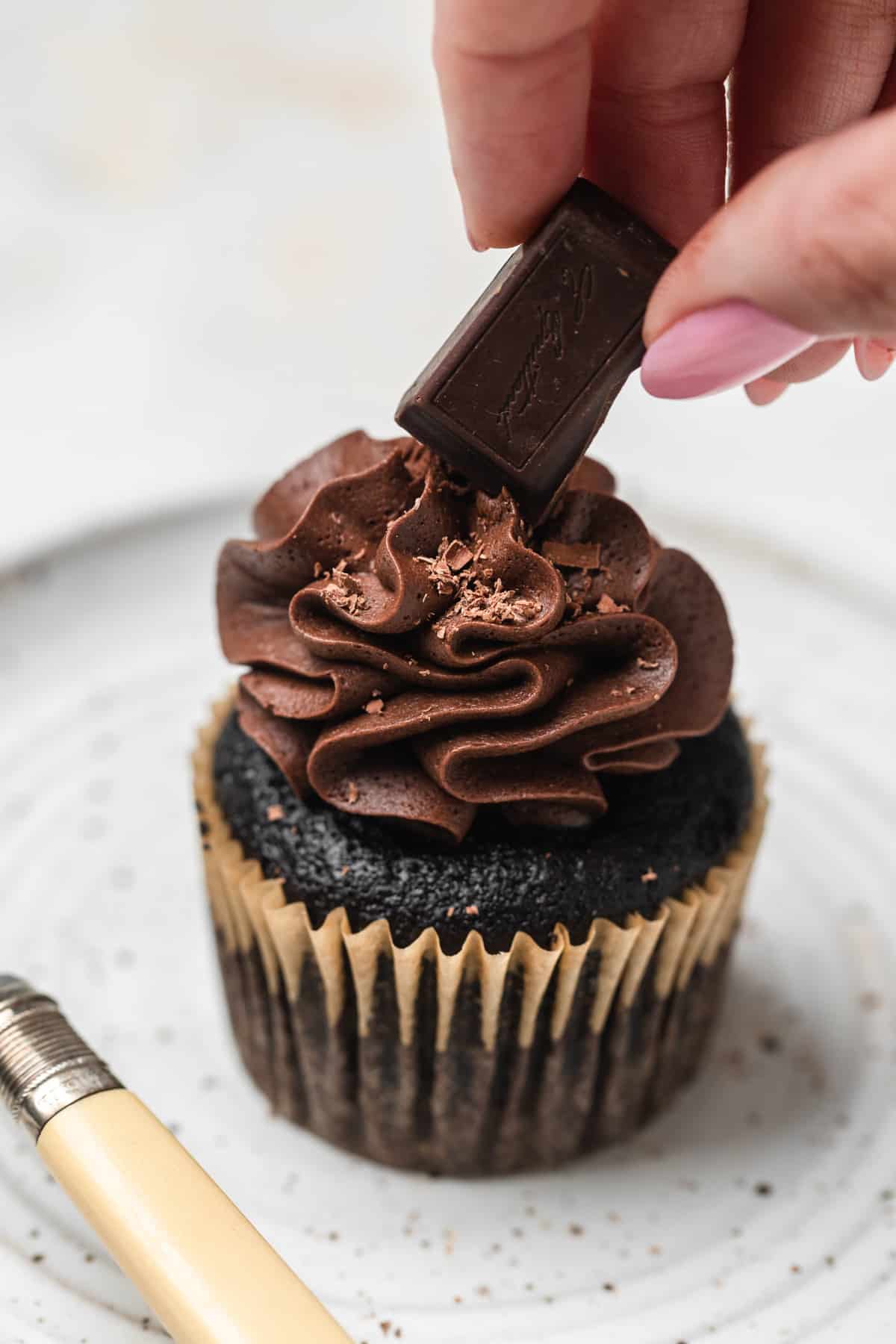 A chocolate cupcake with a piece of chocolate being placed on top.