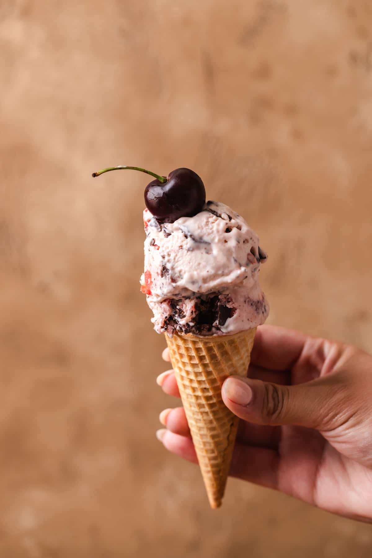 A hand holding up an ice cream cone garnished with a cherry.