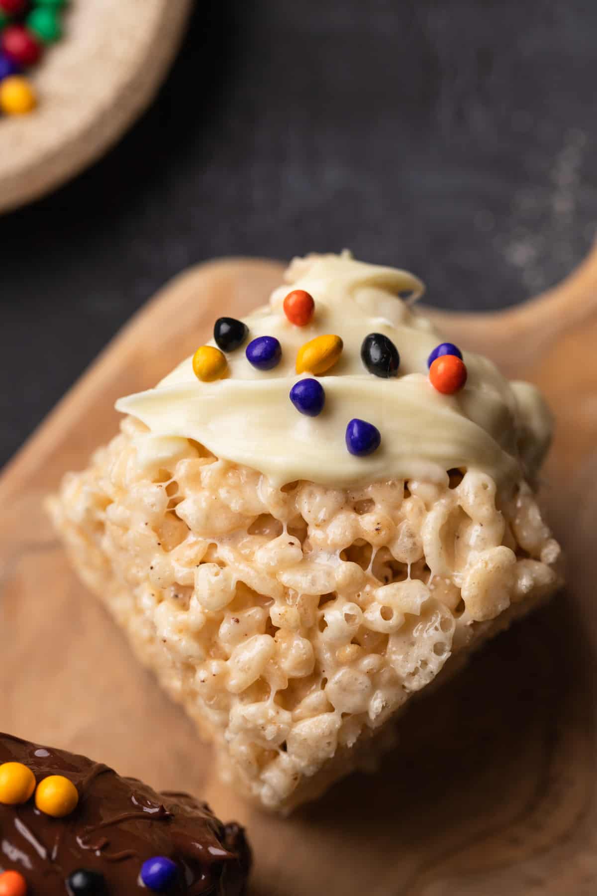 A close up of a Rice Krispie treat with white chocolate and sprinkles on a wooden board.