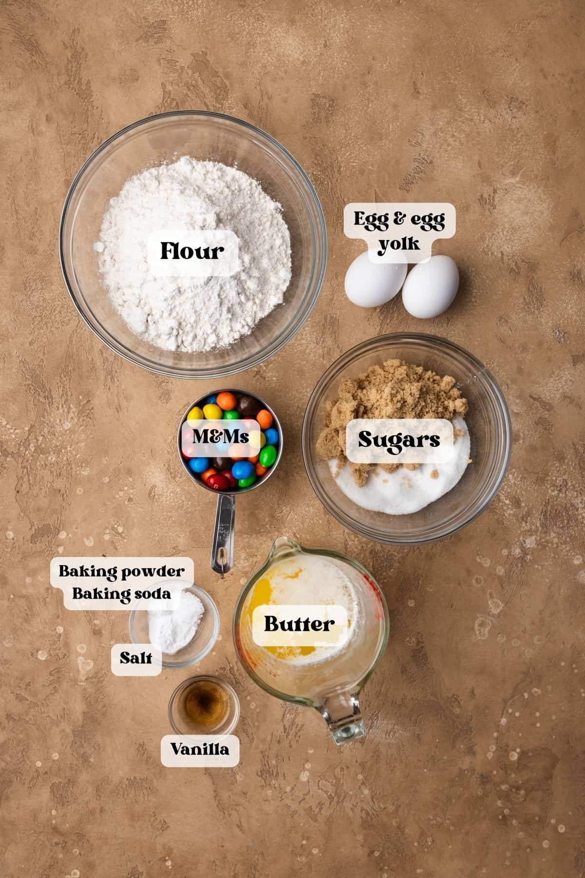 Ingredients to make peanut M&M cookies on a brown surface.