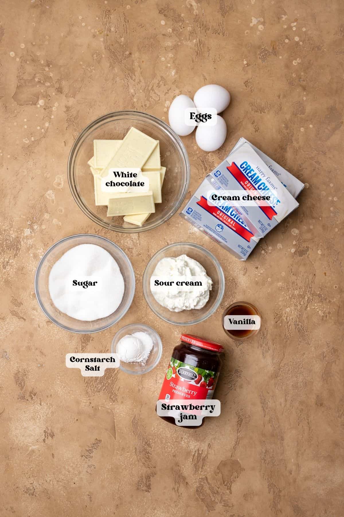 Ingredients to make strawberry white chocolate cheesecake on a tan surface.