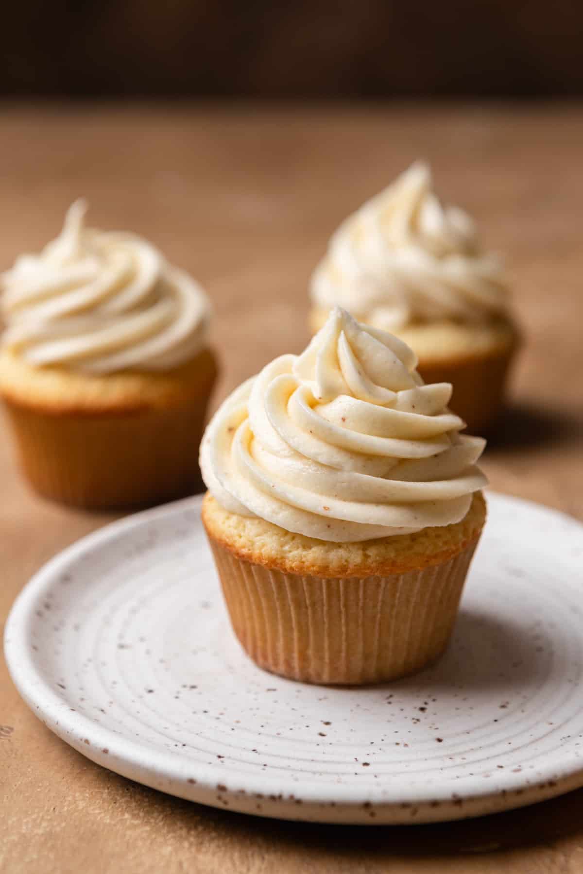 Vanilla cupcakes with oil on a tan surface with one on a white plate.
