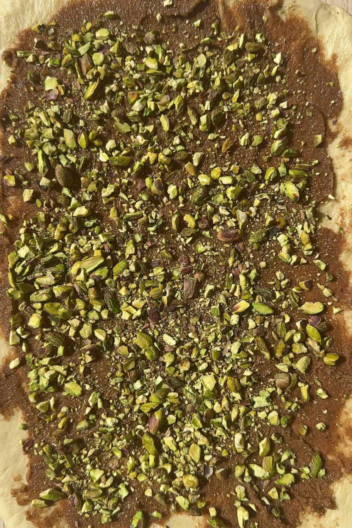 A rectangle of cinnamon roll dough with cinnamon butter and chopped pistachios sprinkled on top.