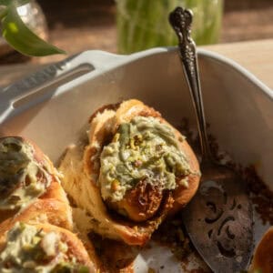 A close up of a pistachio cinnamon roll with pistachio cream cheese frosting in a white pan.