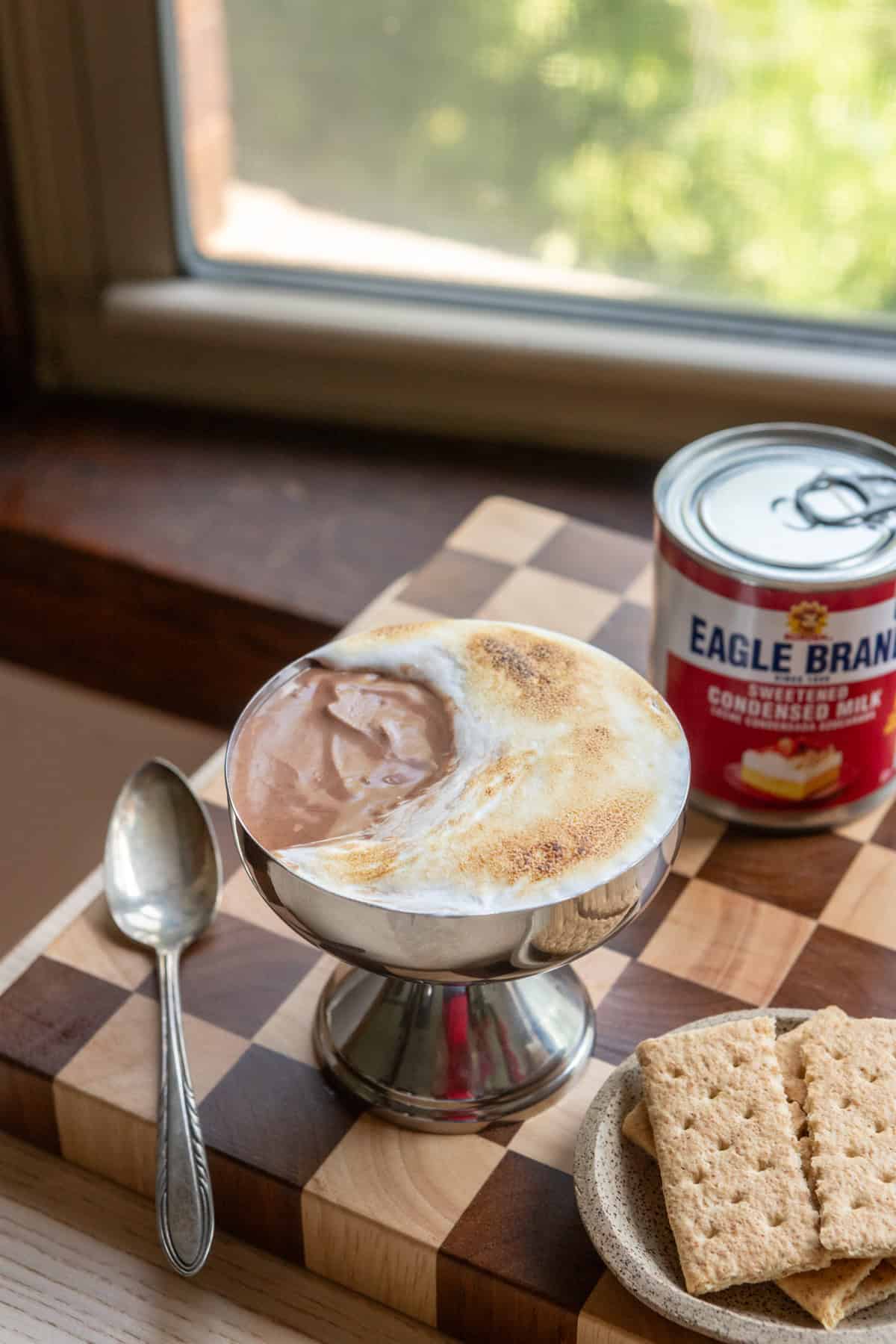 A s'mores cup topped with marshmallow fluff on a checkered cutting board with a spoon.