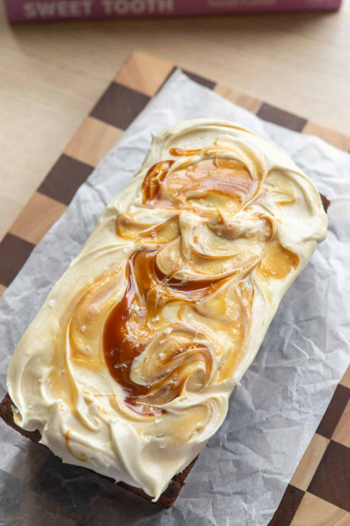 A loaf of frosted salted caramel banana bread on a checkered cutting board lined with parchment paper.
