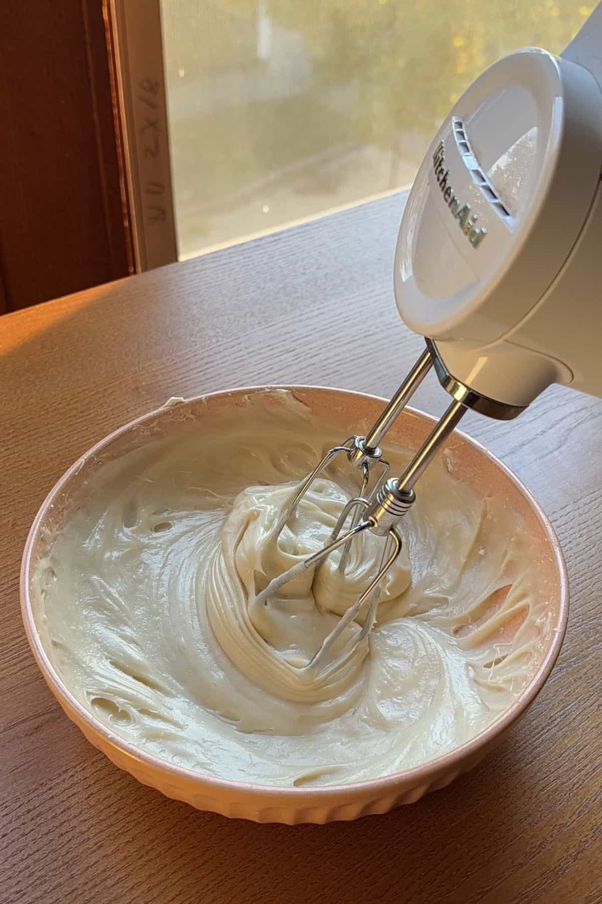 Cream cheese frosting being mixed in a pink bowl with a handheld mixer.